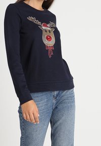 Námořnický modrý sweatshirt s dlouhým rukávem, zdobený grafikou soba s parohy, červeným nosem a mašlí, vyrobený z měkké tkaniny s žebrovanými manžetami a lemem.