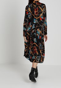 Vestido negro con estampados de cadenas y adornos coloridos, mangas largas, falda plisada y botas negras de tacón alto.