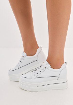 Weiße Plateausneaker mit schwarzem Rand, barfuß getragen, vor einfachem weißem Hintergrund.