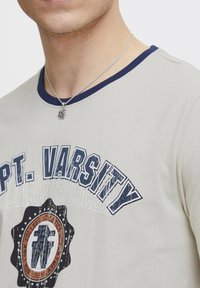 Šviesiai pilka marškinėliai su tamsiai mėlynu apykakle, kuriuose priekyje yra spausdintas tekstas "CPT. VARSITY" ir apvalus grafinis dizainas.