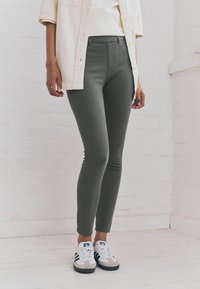 Grijze slim-fit broek van rekbaar materiaal, gecombineerd met witte sneakers met blauwe en zwarte accenten, tegen een licht bakstenen achtergrond.