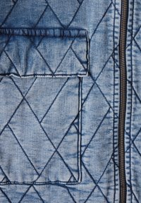 Sininen denimpuku, jossa on viistokuvio, vetoketjukiinnitys ja ommeltu tasku.