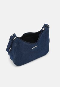 Sac bandoulière en denim bleu foncé avec fermeture éclair, logo argenté, et bandoulière ajustable, présentant une texture douce et une forme arrondie.