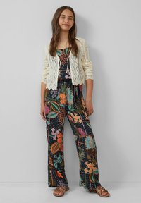 Bloemen jumpsuit met brede benen, donkere achtergrond, meerkleurige patronen; gelaagd met een crèmekleurige, gebreide cardigan met ruitpatronen.