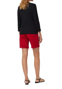 Zwart geribd langesleeve shirt, gecombineerd met rode opgerolde shorts. Natuurlijk haar en platte sandalen maken de outfit compleet.