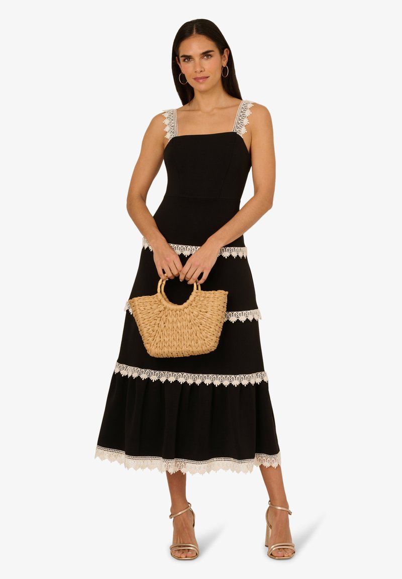 Robe maxi noire avec des bretelles et un ourlet en dentelle, mettant en avant des détails à volants. Le mannequin tient un sac à main en paille tressée. Des sandales complètent le look.