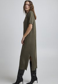 Robe t-shirt longue vert olive en matériau doux, avec des manches courtes, des fentes sur les côtés et une coupe décontractée, associée à des bottes noires jusqu'aux genoux.