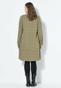 Olivegrünes karierter Kleid mit langen Ärmeln, einem gerafften Saum und einem lockeren Schnitt. Getragen über schwarzen Leggings und Stiefeletten.
