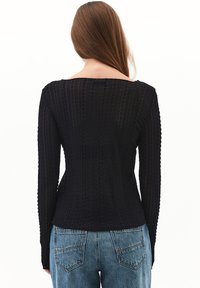 Kvinde med langt brunt hår iført en struktureret sort langærmet top og blå jeans, vist fra ryggen mod en hvid baggrund.