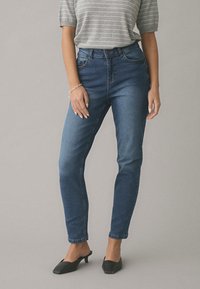 Žena oblečená v modrých džínsoch s vysokým pásom, sivom pruhovanom tričku s krátkym rukávom, čiernych topánkach s nízkym podpätkom a striebornom náramku na ľavom zápästí.