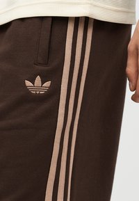 Nahaufnahme brauner Hose mit beigen drei Streifen und Adidas-Trefoil-Logo, getragen zu einem beigen gerippten Top, und einer Hand an der Seite.