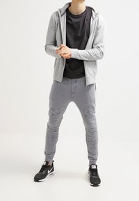 Šedá zipová mikina přes černé tričko, doplněná šedými tepláky s žebrovanými akcenty a černými teniskami s bílými detaily.