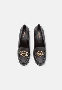 Mocassins en cuir noir avec un bout arrondi, présentant un détail en métal doré en forme de cercle et une finition lisse. Accentué de coutures le long des bordures.