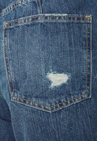 El bolsillo trasero de unos jeans de denim azul con tela blanca desgastada y deshilachada asomando a través de un pequeño desgarro cerca de la esquina inferior derecha.
