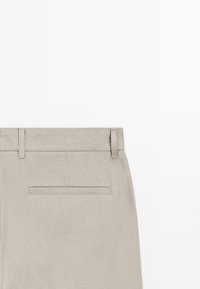 Pantalon beige clair avec passants pour ceinture et une unique poche arrière horizontale sur un fond blanc uni.