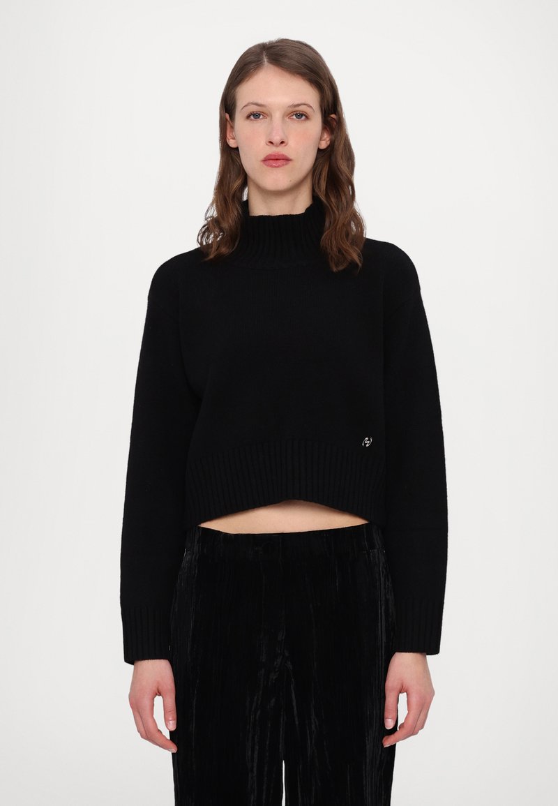 Schwarzer, kurzer Pullover mit geripptem Kragen und Bündchen. Weicher Strickstoff. Kombiniert mit schwarzen, plissierten Hosen, die einen monochromen Look schaffen.