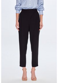 Femme portant un pantalon noir ajusté taille haute, des talons nus à bande de cheville et une chemise à rayures bleues avec les manches retroussées.