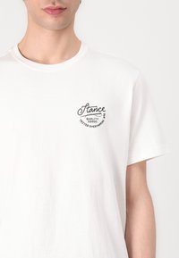 Weißes Baumwoll-T-Shirt mit rundem Ausschnitt und kurzen Ärmeln. Mit einem schwarzen Aufdruck auf der linken Brustseite. Weicher Stoff.