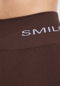 Leggings marroni con una vita a costine che presenta il testo bianco "SMIL". Il tessuto appare liscio ed elastico, con un design a vestibilità aderente.