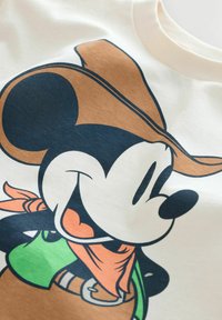 Bomulds t-shirt med en tegneseriefigur iført en brun cowboyhat, orange bandana, grøn skjorte og beige baggrund.