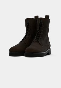 Bottines en suède marron avec un bout arrondi, un laçage à l'avant et une fermeture éclair sur le côté. Semelle extérieure plate noire avec une texture antidérapante.