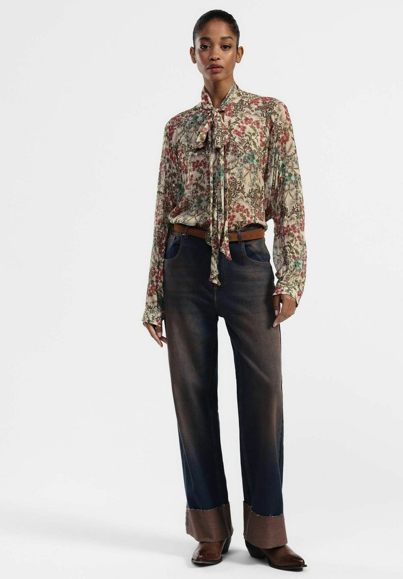 Blusa floreale con collo a cravatta, caratterizzata da motivi crema, rossi e blu. Pantaloni larghi in denim scuro con risvolti marroni a contrasto e stivali.