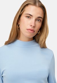 Haut bleu clair à manches longues avec un col montant. Tissu lisse, coupe ajustée. Boucles d'oreilles anneaux dorés remarquables. Portrait en gros plan.