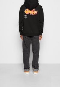Czarna hoodie z dużym pomarańczowym logo w gradiencie z tyłu, w połączeniu z ciemnymi dżinsami i trampkami z pomarańczowymi akcentami.