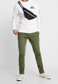 Svart nylon midjeväska med vit rem, bärs över en vit långärmad skjorta och olivgröna byxor; visas med neutrala sneakers.