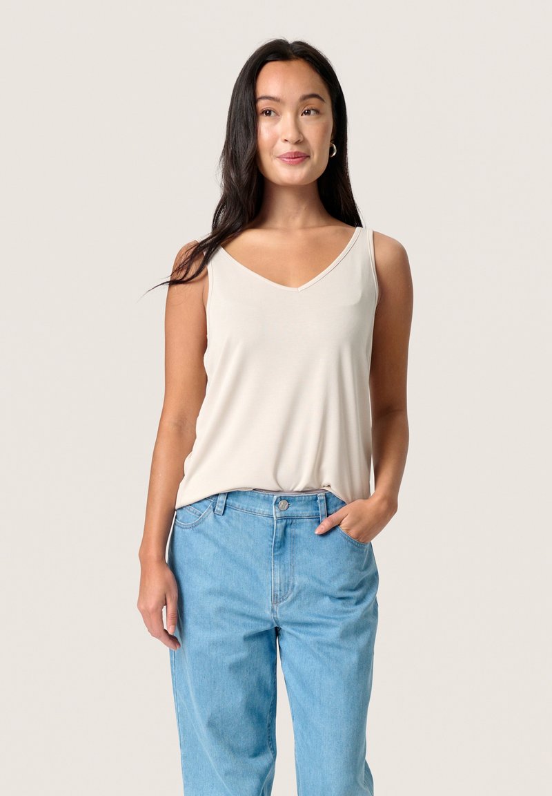 Beige sleeveless top with a V-neck, made from soft fabric, draping slightly, paired with high-waisted light blue denim jeans.