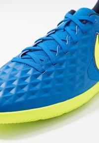 Zapatilla deportiva azul con una superficie texturizada que presenta patrones triangulares y una suela de color amarillo neón. Cordones y elementos de malla en azul a juego.