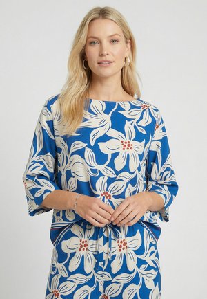 Femme blonde en haut bleu et pantalon avec un grand imprimé floral blanc et des accents rouges, debout devant un fond gris clair.