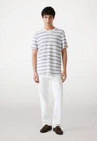 Camiseta de punto a rayas en blanco con rayas azul claro y azul oscuro, acompañada de pantalones blancos y zapatos estilo mocasín marrones.