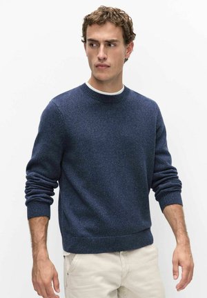 Jonge man met een blauw gebreide trui en lichtgekleurde broek, staand met ontspannen handen langs zijn zij, kijkt naar rechts tegen een witte achtergrond.