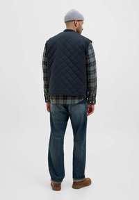Gilet trapuntato blu navy senza maniche, indossato sopra una camicia a quadri verde, abbinato a jeans blu e scarpe marroni, visto da dietro.