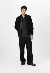 Juoda suede bomber striukė su kontrastingu tekstūruotu užtrauktuku ir apykakle, derinama su plačiomis juodomis kelnėmis ir blizgiais juodais batais.