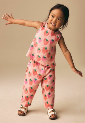 Ung pige i lyserød top og bukser med jordbærprint, balancerer med armene udstrakt, smilende, mod en ensfarvet beige baggrund.