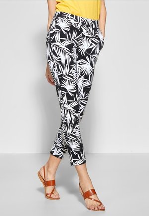 Persoon die zwarte en witte tropical print cropped broek draagt, gele top en bruine sandalen met bandjes en platte zolen, staand tegen een effen achtergrond.