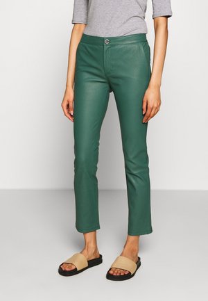 Woman wearing green leather straight-leg pants, gray three-quarter sleeve top, and beige slide sandals standing on a gray floor.