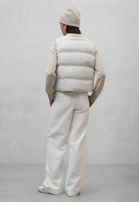 Chaleco plumas blanco con cuello alto, usado sobre un suéter beis claro. Combinado con pantalones anchos blancos y zapatos beige, en un fondo neutro.