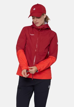 Vrouw die de mouw van een tweekleurige rode outdoorjas aanpast, met een bijpassende rode pet en zwarte broek tegen een effen witte achtergrond.