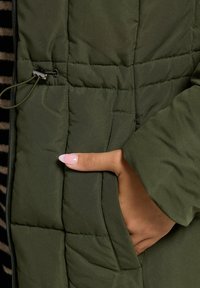 Grön quiltad puffjacka med sidofickor, dragkedjedetaljer och en slät textur. Tyget verkar hållbart och väderbeständigt.