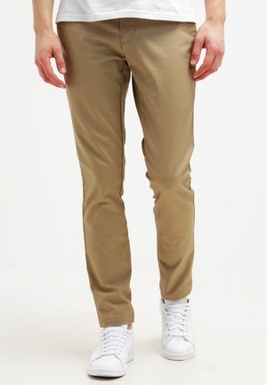 Mand iført tætsiddende beige bukser med lommer, hvide sneakers og en hvid skjorte, stående mod en ensartet lys baggrund.