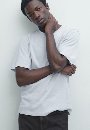 Giovane uomo con indosso una maglietta grigio chiaro e pantaloni scuri, in piedi con una mano sul collo e l'altra incrociata sul torso.