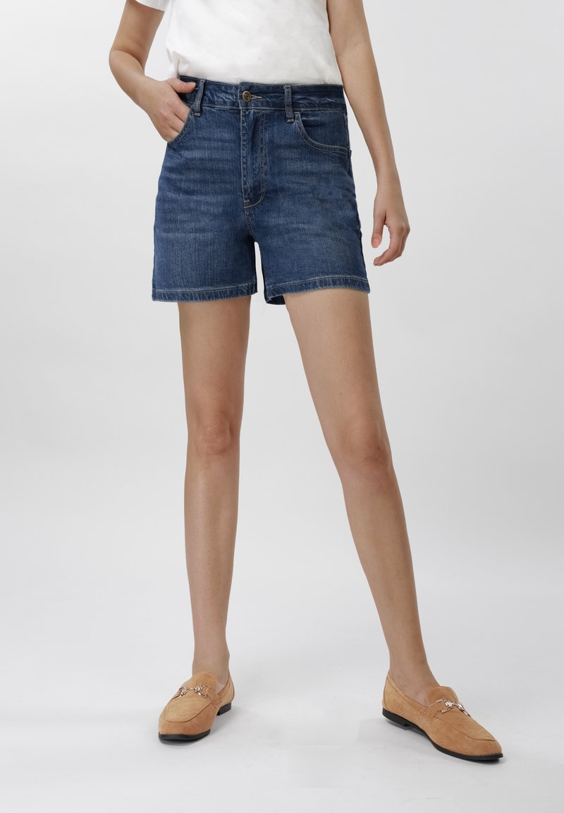 Person iført mørkeblå denimshorts, hvit skjorte og brune loafers, stående med en hånd i lommen mot en ensfarget bakgrunn.