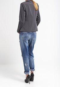 Kobieta wearing an dark gray blazer, distressed rolled-up blue jeans, and black high-heeled shoes, standing with hands in pockets, facing away.