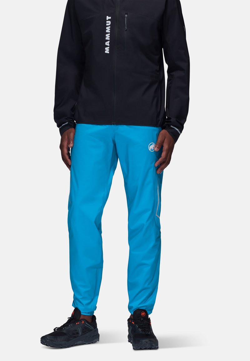 Svart softshell-jacka med vitt logotyp, tillsammans med blåa vattentäta byxor med elastiska manschetter. Svarta skor med röda detaljer fulländar outfiten.