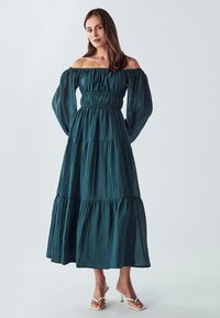 Haljina u boji tirkiznog zelena s bretelama, slojevitom tkaninom, elastičnim pojasom, dugim lepršavim rukavima i opuštenim krojem, uparena s bijelim sandalama na petu.