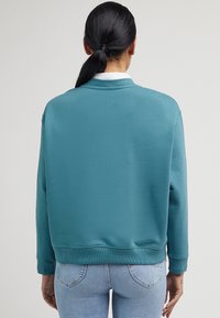 Sudadera de color verde azulado con un corte holgado, cuello, puños y dobladillo de canalé. Está fabricada con un tejido suave, presenta un diseño sencillo y sin patrones visibles.
