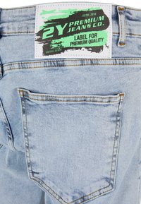 Világoskék farmernadrág hátsó zsebbel, amelyen egy fehér címke látható, zöld és fekete grafikával, amelyen az "2Y PREMIUM JEANS CO." és a "LABEL FOR PREMIUM QUALITY." felirat olvasható.